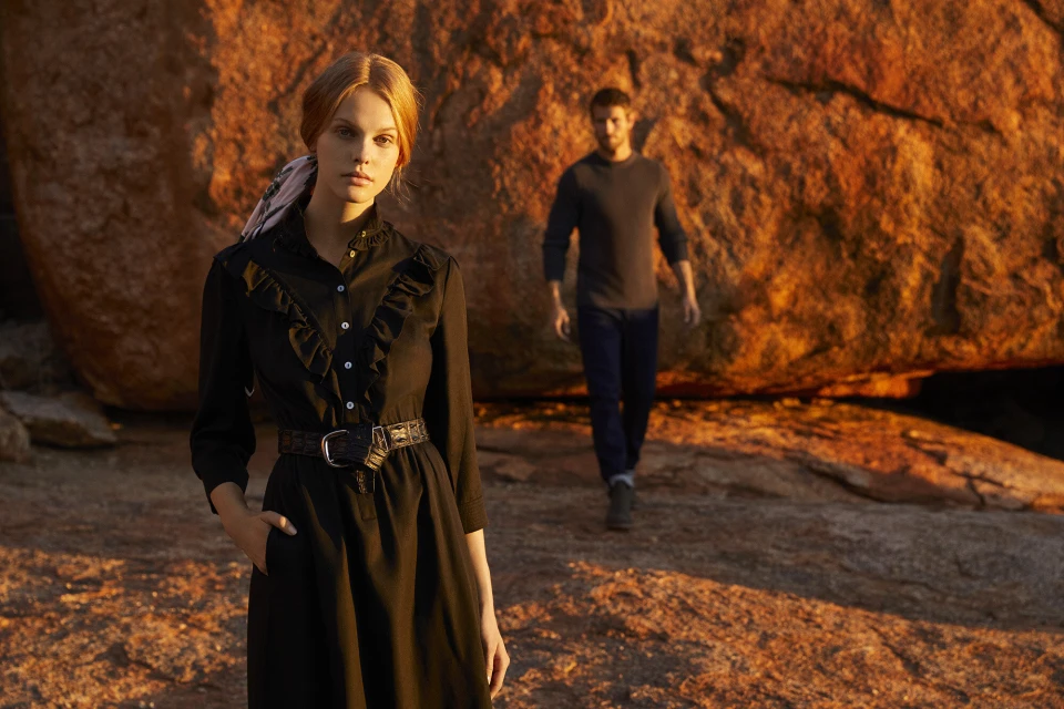 A woman in a black dress stands in the foreground as a man walks in the background on a rocky, orange terrain under the warm sunlight.