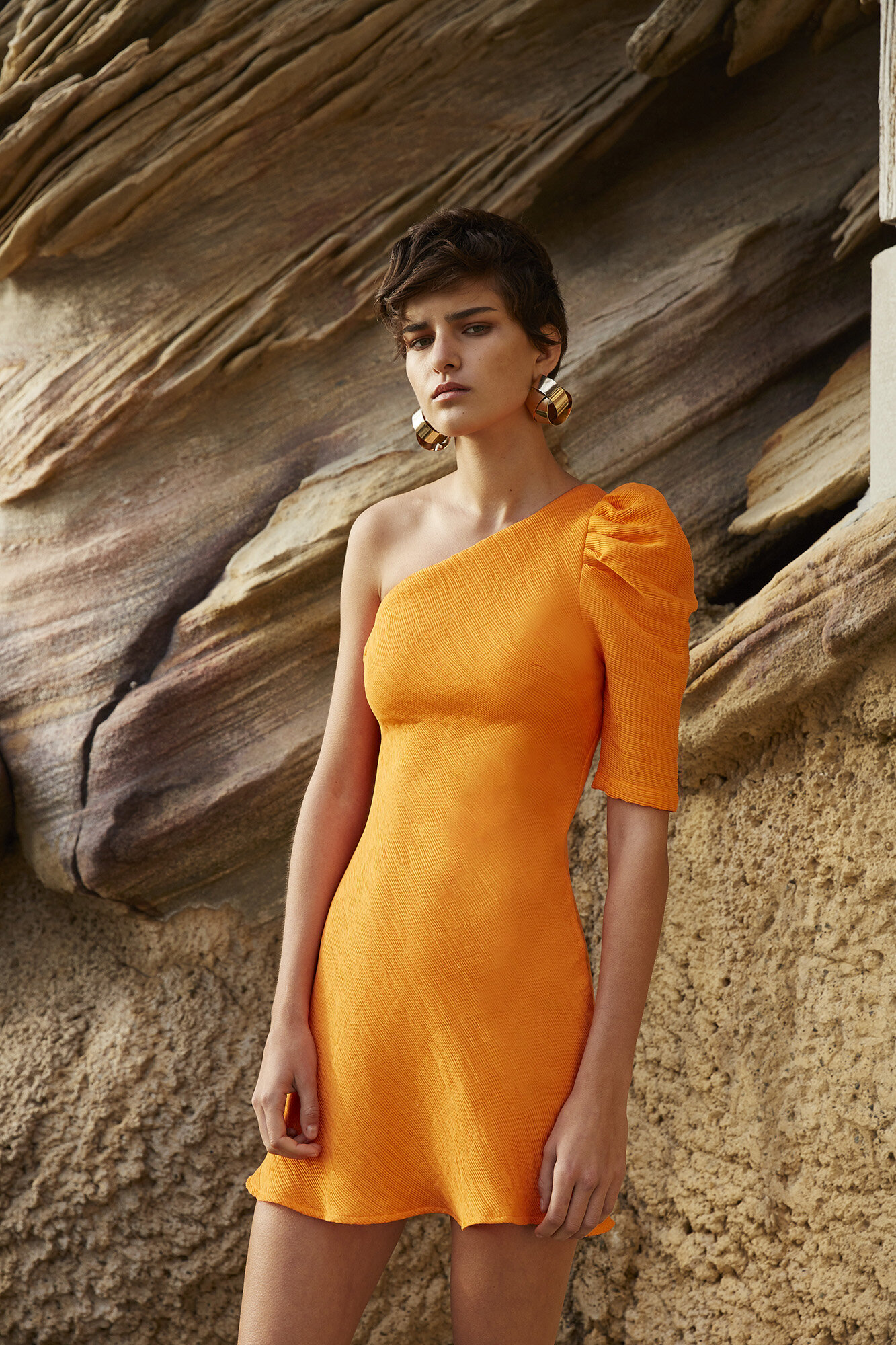 Adult female poses in a short, orange, one-shoulder dress against a rocky background, accessorized with large, hoop earrings. Fashion shot.