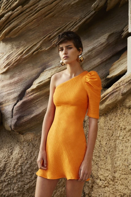 Adult female poses in a short, orange, one-shoulder dress against a rocky background, accessorized with large, hoop earrings. Fashion shot.