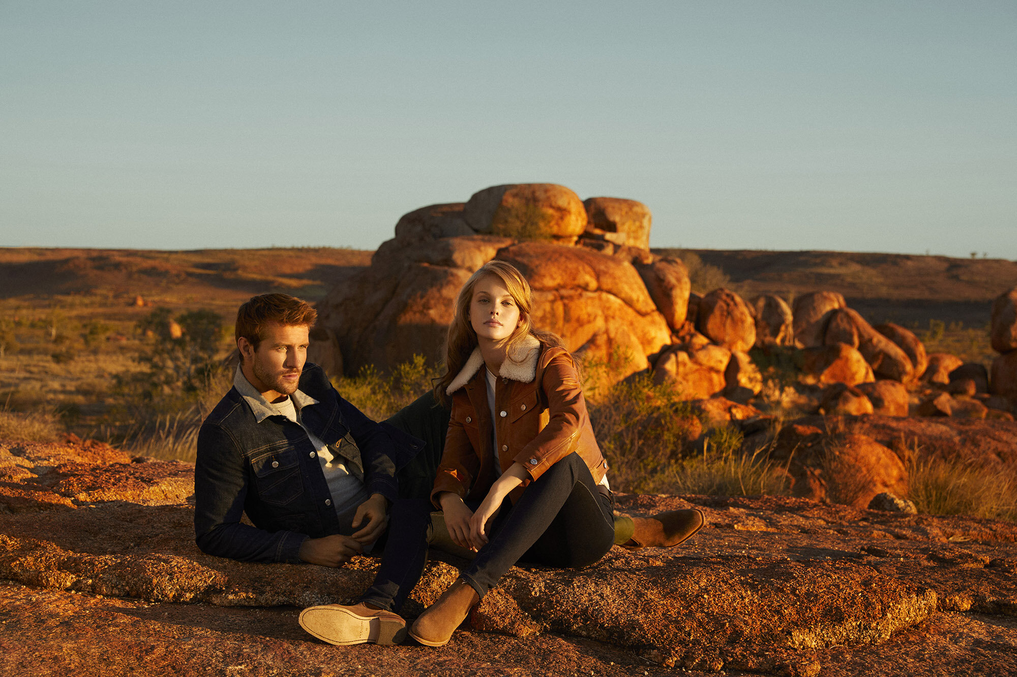 A couple models denim jackets and suede shoes while sitting on rocks in an outdoor desert environment during golden hour.