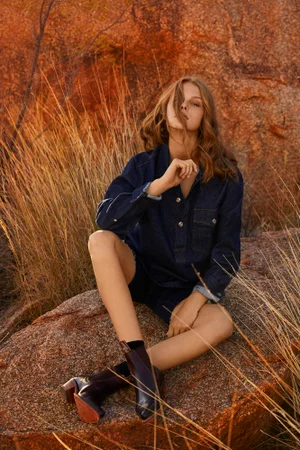Young woman in a denim shirt and dark boots seated on a large red rock surrounded by dry grass.