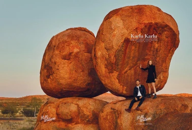 A man and woman posing among massive rust-colored granite boulders in an arid outback landscape at sunset.
