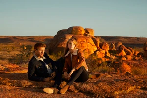 Man and woman seated on desert rocks at sunset, wearing denim and a shearling jacket, photographed by Daniella Rose in Sydney.