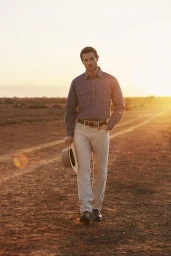 Man walking on a dirt road at sunset wearing a blue work shirt and light trousers while holding a hat