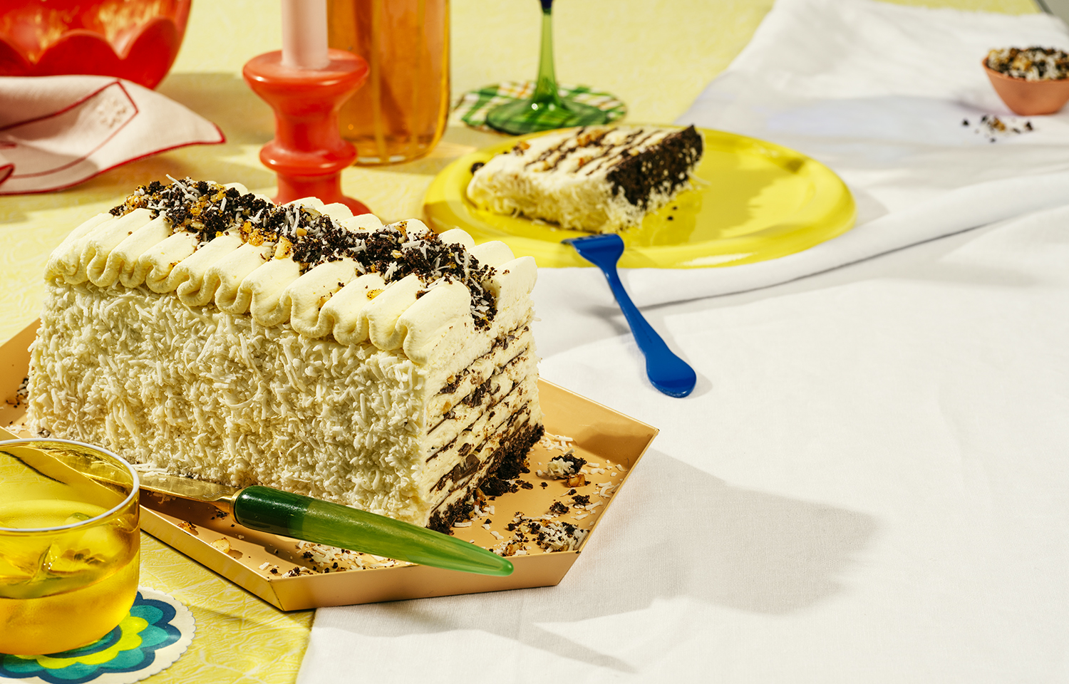 A slice of multi-layered chocolate cake with white frosting and toppings sits on a table with a plate, drink, and candle.