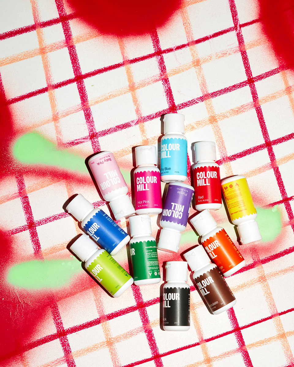 Multiple small bottles of colourful food dye arranged on a white surface with a pastel spray-paint style grid pattern in red, orange, and green.