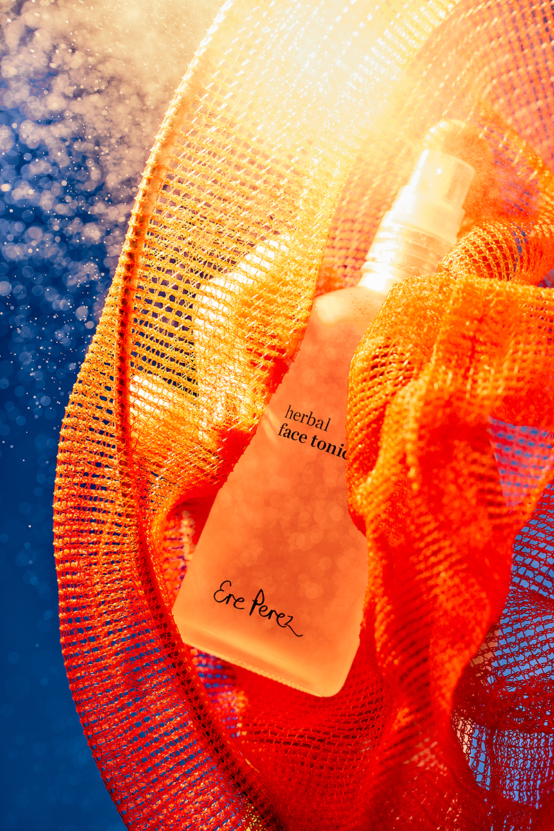 Ere Perez herbal face tonic, in frosted bottle, surrounded by an orange mesh, against a blue background, illuminated by a bright light.