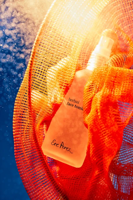 Ere Perez herbal face tonic, in frosted bottle, surrounded by an orange mesh, against a blue background, illuminated by a bright light.