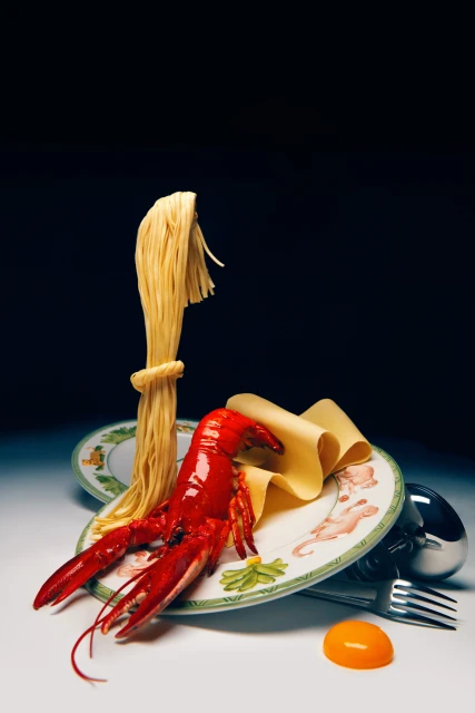 Stylized food photography of cooked pasta, a red lobster, and a raw egg yolk served on decorative plates against a dark backdrop.