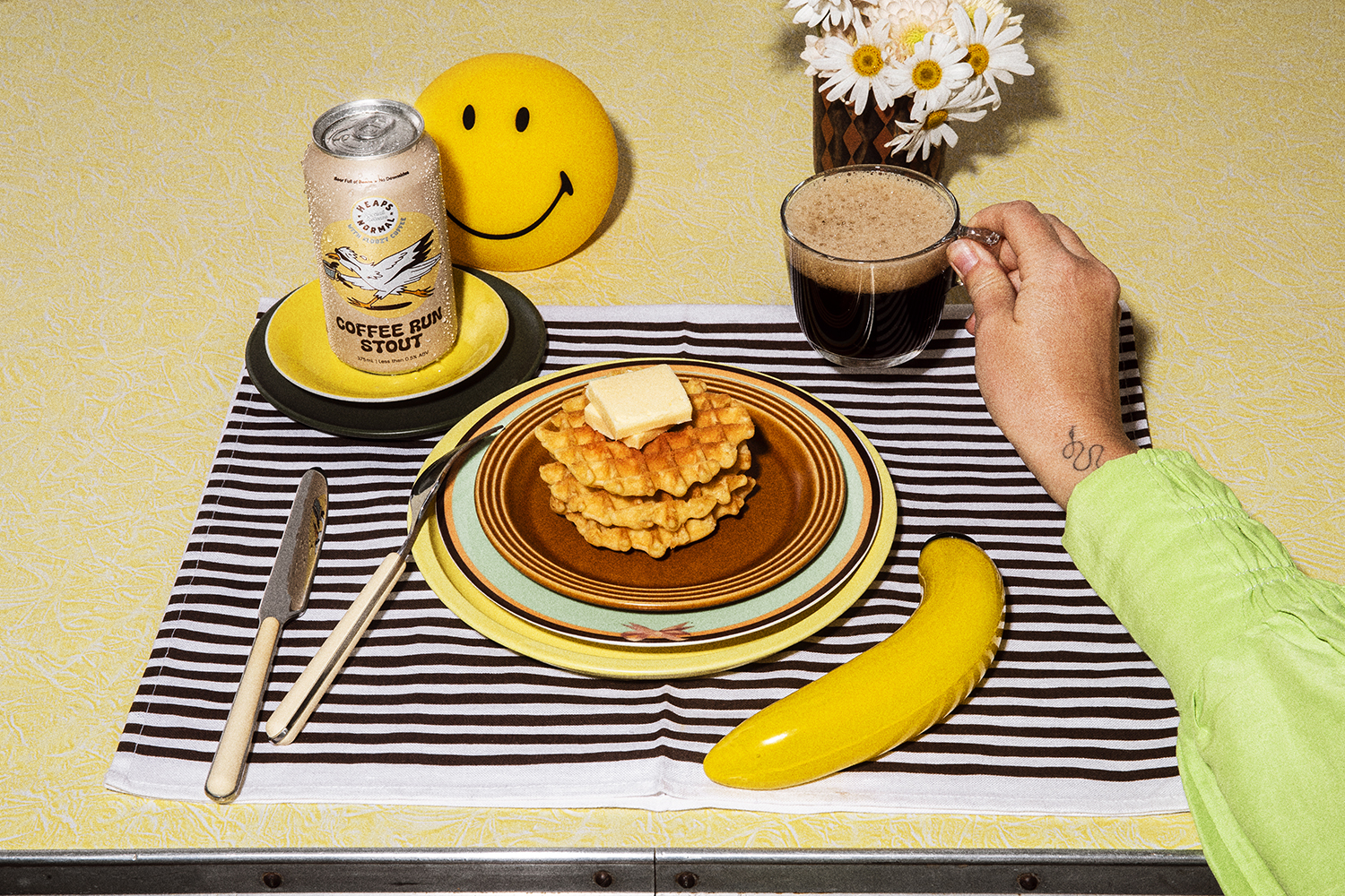 Overhead shot of waffles with butter, a can of stout, a mug of coffee, and a smiley face decoration on a patterned tablecloth.