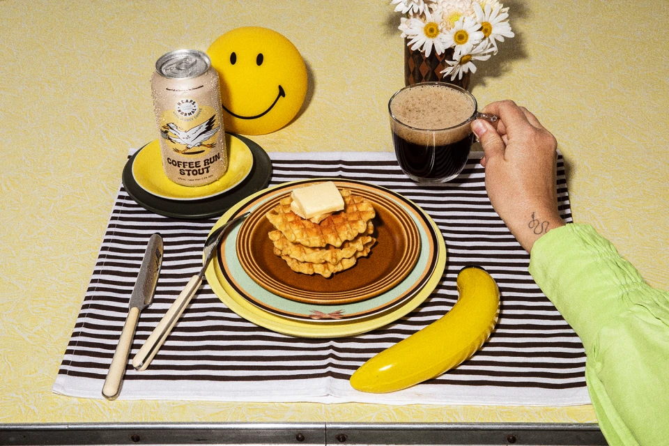 Overhead shot of waffles with butter, a can of stout, a mug of coffee, and a smiley face decoration on a patterned tablecloth.