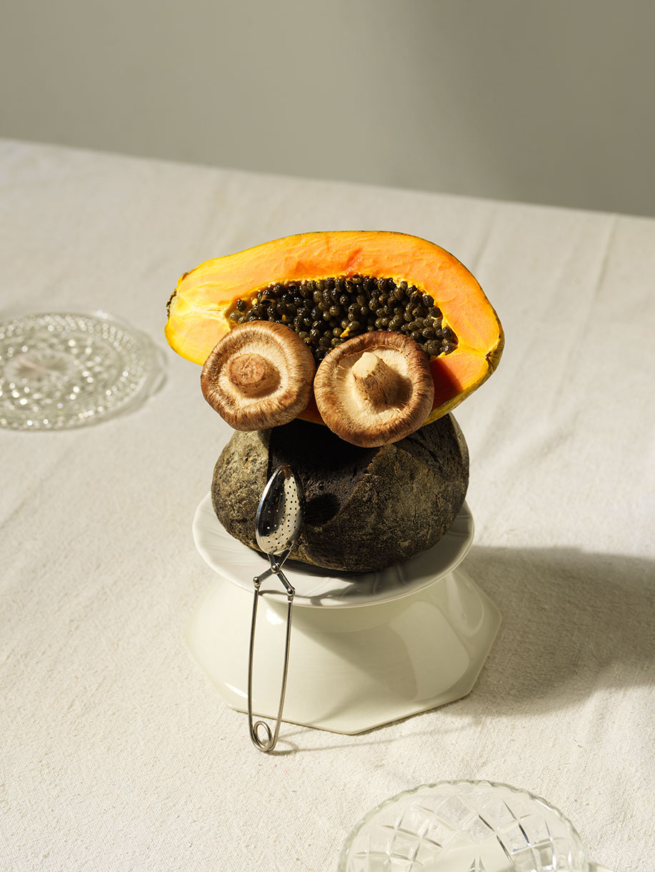 Still life of papaya, mushrooms, rock, and tea infuser arranged as a face on a table with neutral background and natural lighting.