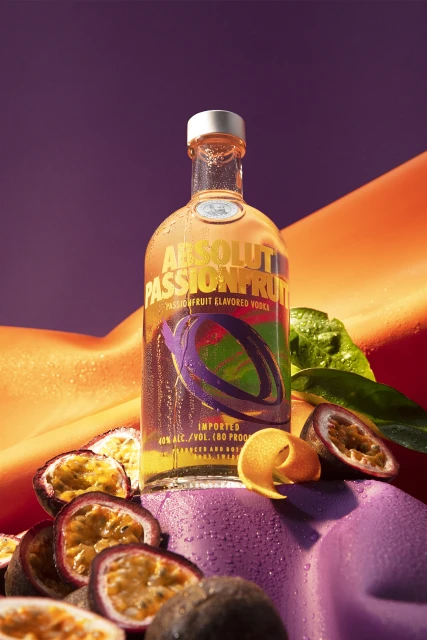 Absolut Passionfruit vodka bottle on purple surface with sliced passion fruit, orange peel, and a purple and orange backdrop.