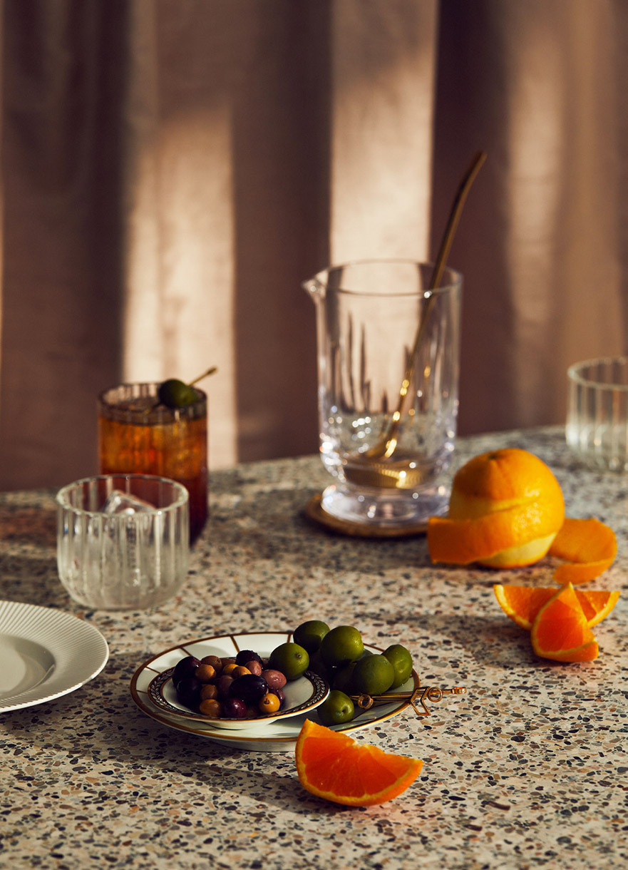 Still life shot of an orange next to olives, a cocktail, and an empty glass on a speckled table, set against a neutral curtain backdrop.