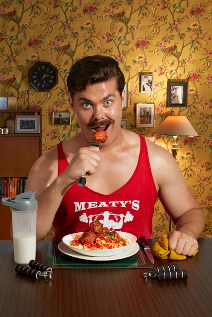 Weightlifter eats a meatball on a fork at a table with spaghetti, free weights, and a protein shake in a vintage-styled dining room.
