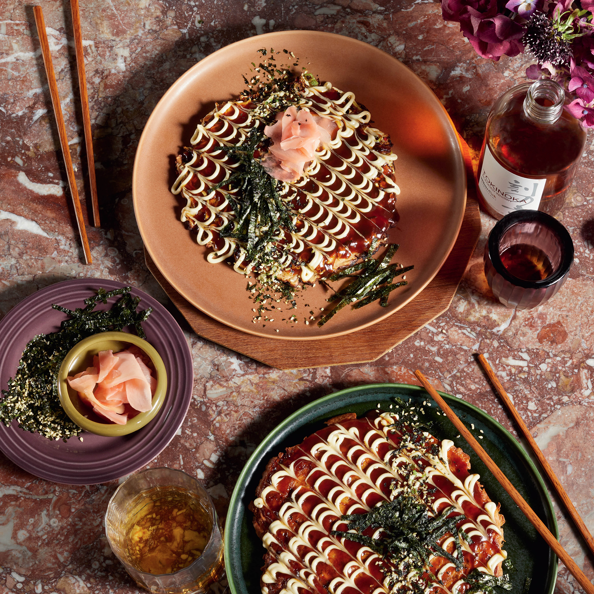Overhead shot of okonomiyaki dishes with toppings and drinks on a marble table, styled with chopsticks and purple flowers, inviting to eat.