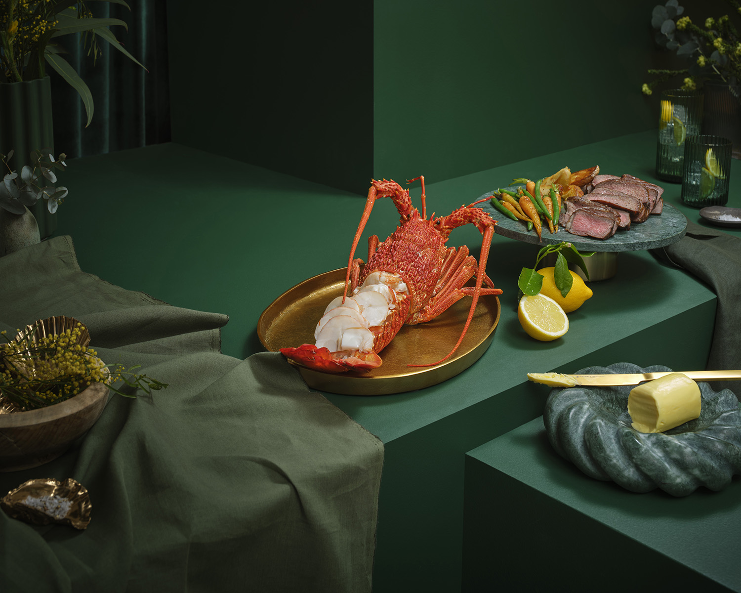 Still life food photography of a spiny lobster with sliced tail meat on a golden plate, accompanied by sliced steak, lemon, and butter.