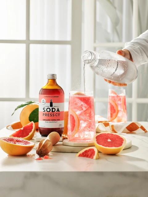Pouring soda water into a glass with pink grapefruit slices, next to a Soda Press Co. bottle and marble surface, and a window backdrop.