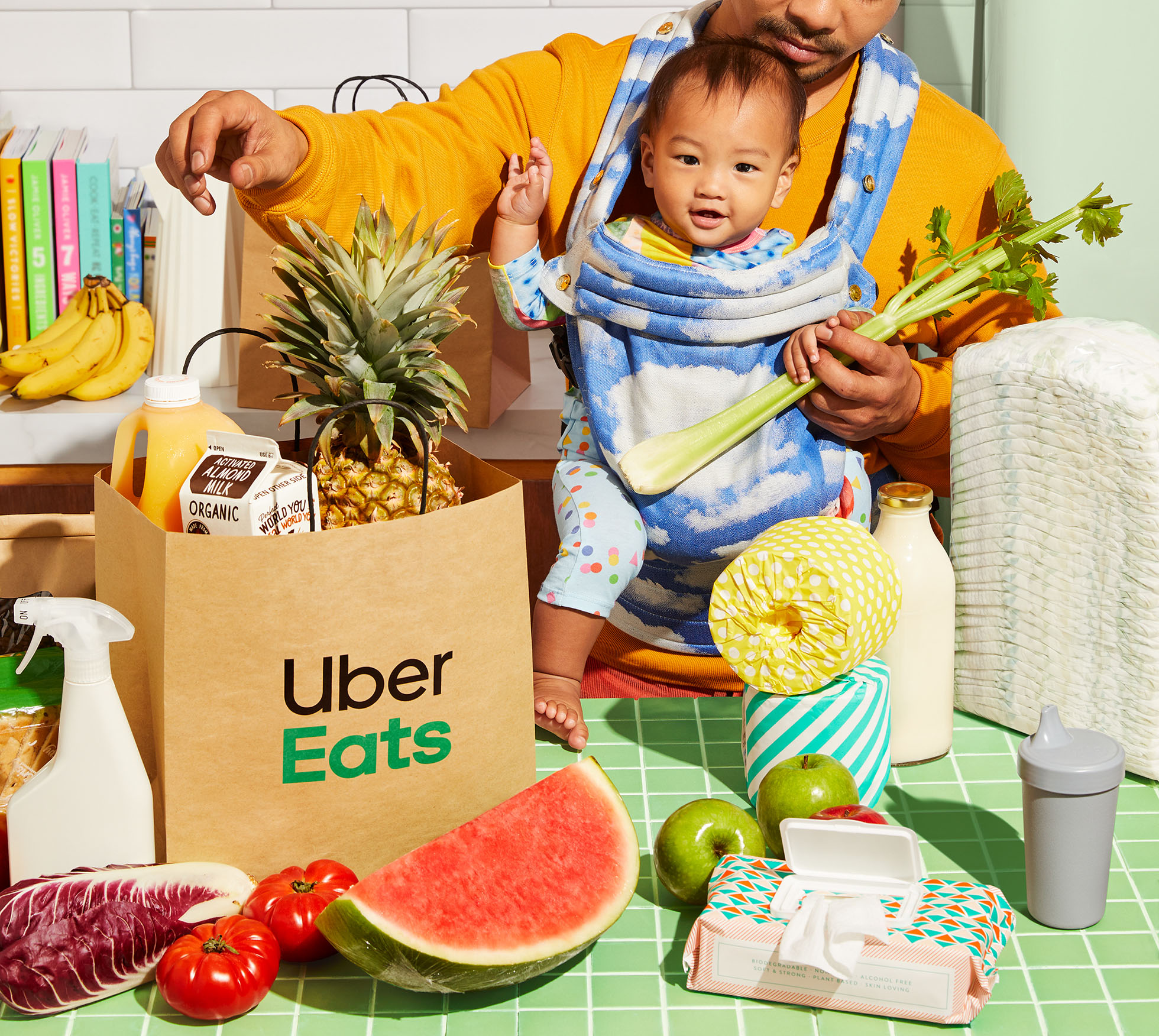Adult carries a baby in a carrier with a grocery bag full of pineapple, juice, and produce on a green checkered surface.