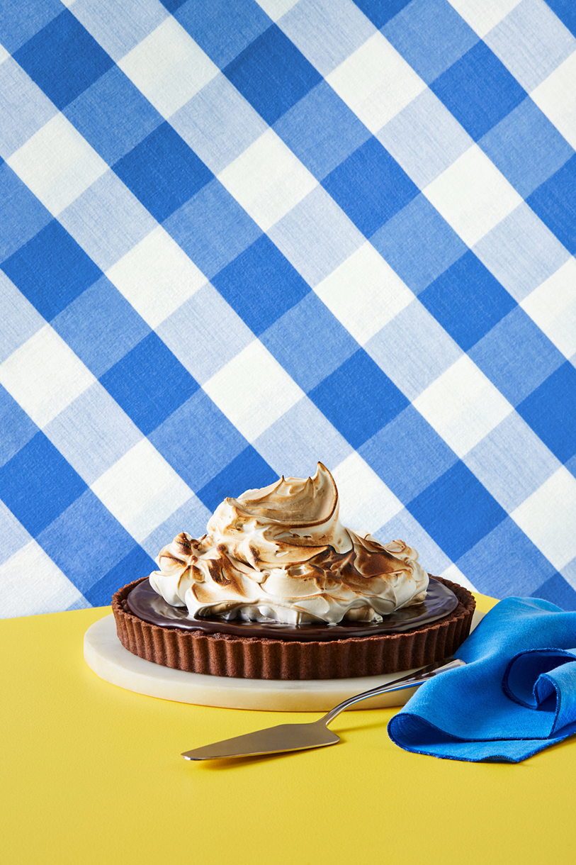 Overhead shot of a chocolate tart with toasted meringue topping on a yellow surface with blue and white checkered backdrop.