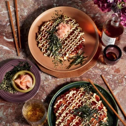 Overhead food styling shot of two okonomiyaki dishes on ceramic plates with chopsticks and glass bottles on a marble table.