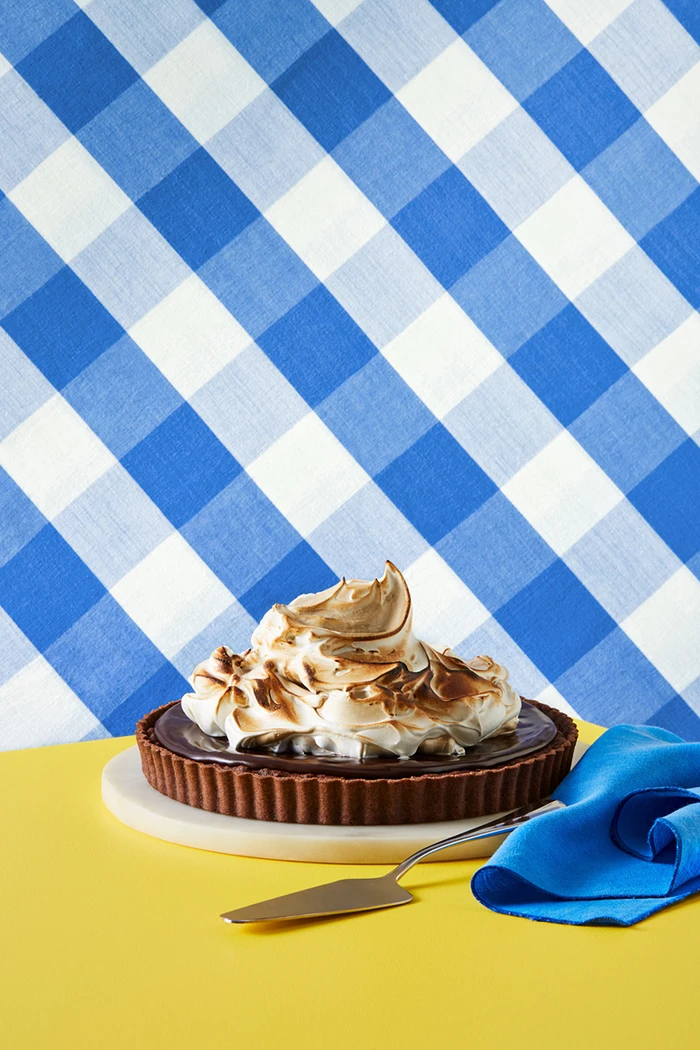 Chocolate tart with torched meringue on a yellow table with a blue napkin and blue gingham background