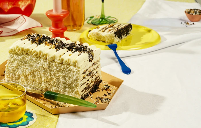 Layered frosted cake garnished with cookie crumbs and coconut flakes on a styled table with colorful glassware