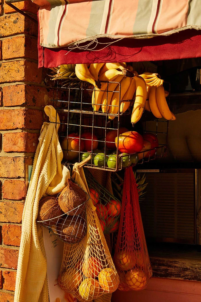 A wire basket holding bananas and mangos, hanging below a striped awning alongside coconuts in a net bag and oranges in a red string bag