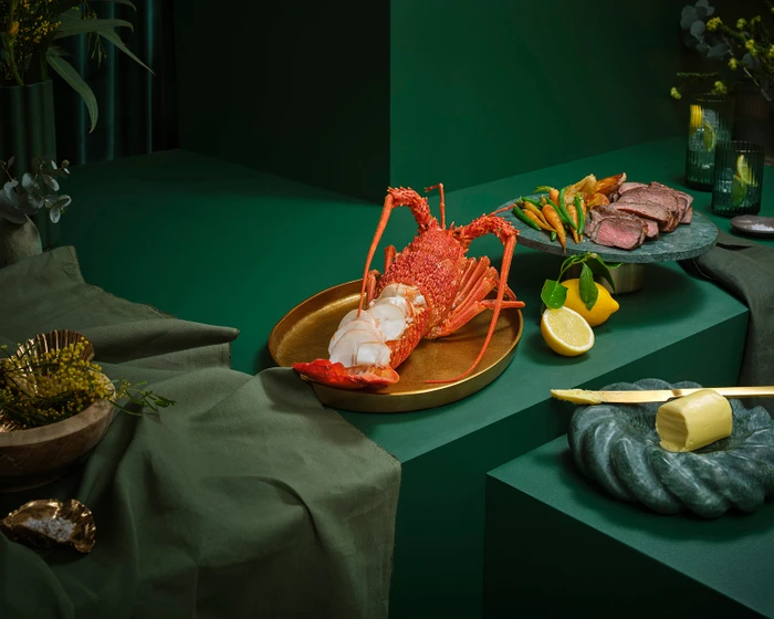 A prepared lobster on a gold platter alongside sliced steak, vegetables, and butter with dark green styling.