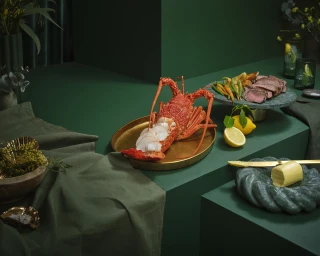 A prepared lobster on a gold platter alongside sliced steak, vegetables, and butter with dark green styling.