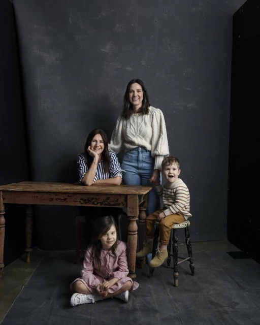 Two adult women, a young girl and boy pose in front of a dark wall near a rustic wooden table and stool in a studio setting.