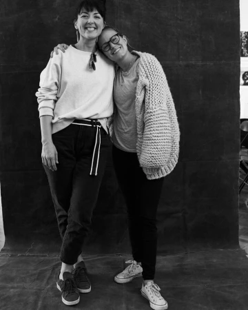 Black and white portrait of two smiling women posing together against a dark textured backdrop by Hugh Stewart
