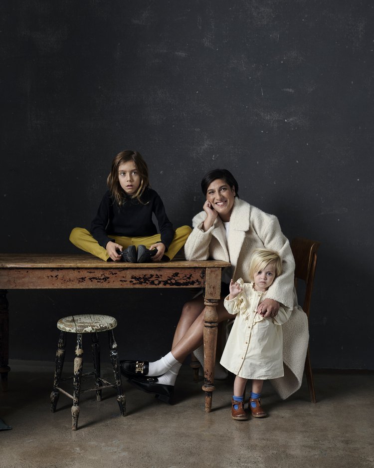Indoor shot of a woman in a white coat with two children, one sitting on a wooden table, and the other standing beside her.
