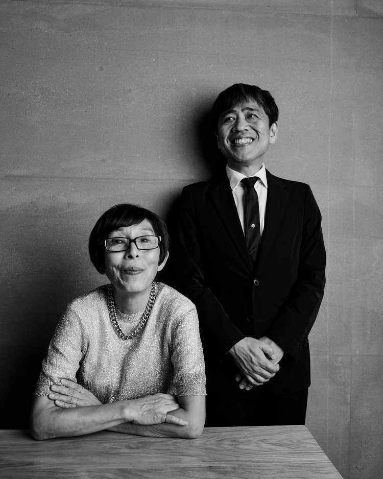 Portrait of an elderly woman seated at a wooden table next to a smiling man in a suit against a textured grey wall