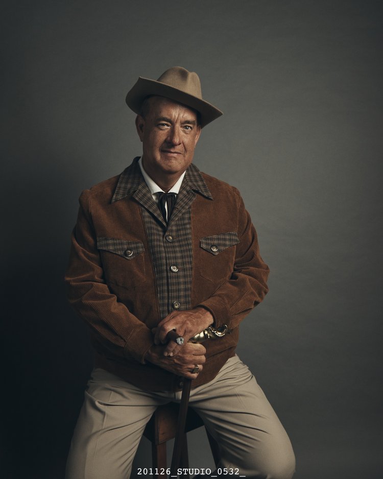 Older man wearing a brown hat and jacket sitting on a chair in a studio holding a cane with both hands looking at the camera.