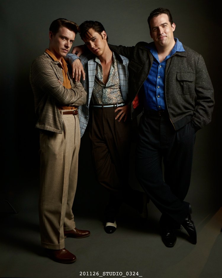 Three men pose in a studio setting, wearing retro-style clothing including suit jackets, button-down shirts, and dress pants, against a grey backdrop.