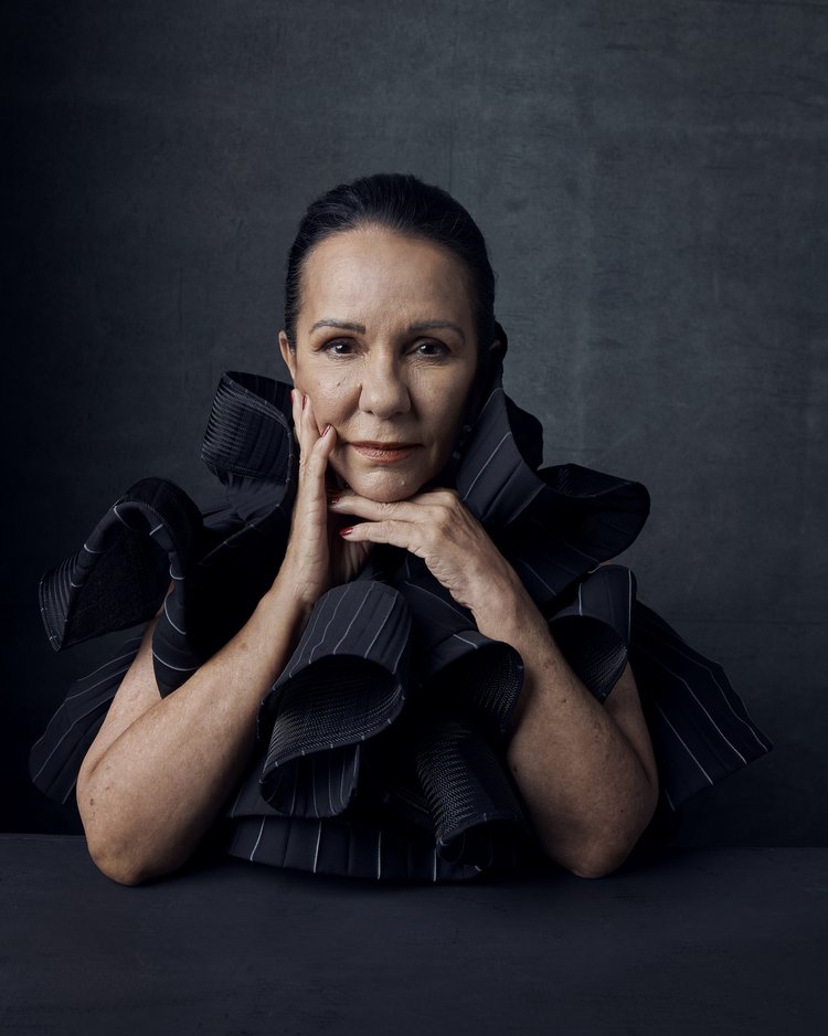 Portrait of an adult woman with hands near her face, wearing a ruffled black and white pinstripe collar against a dark gray background.