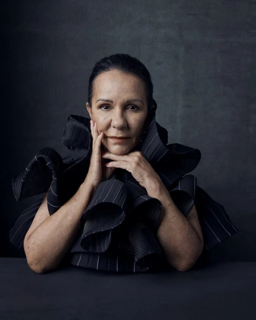 Portrait of an adult woman with hands near her face, wearing a ruffled black and white pinstripe collar against a dark gray background.