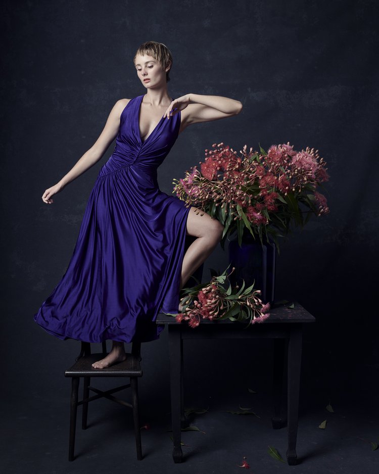 Woman in a purple gown posing next to a flower arrangement on a table in a studio setting with a dark background and soft lighting.