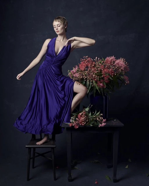 Woman in a purple gown posing next to a flower arrangement on a table in a studio setting with a dark background and soft lighting.
