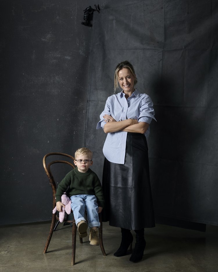 Woman and child in a photo studio setting. The child is seated on a chair, holding a pink stuffed animal, while the adult stands behind.