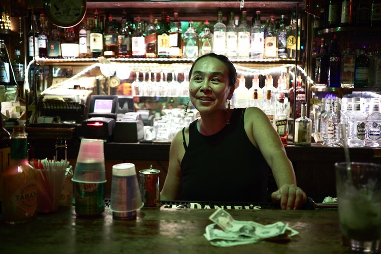 A bartender smiles while standing behind a bar stocked with liquor bottles, glasses, and bar equipment in a dimly lit establishment.
