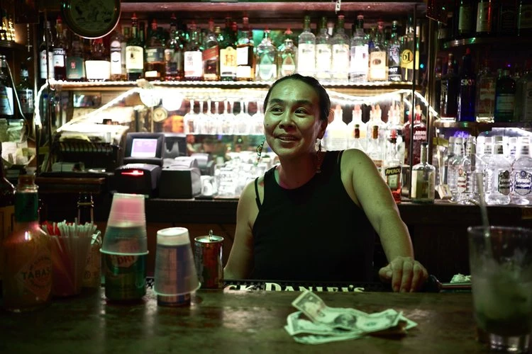 A bartender smiles while standing behind a bar stocked with liquor bottles, glasses, and bar equipment in a dimly lit establishment.