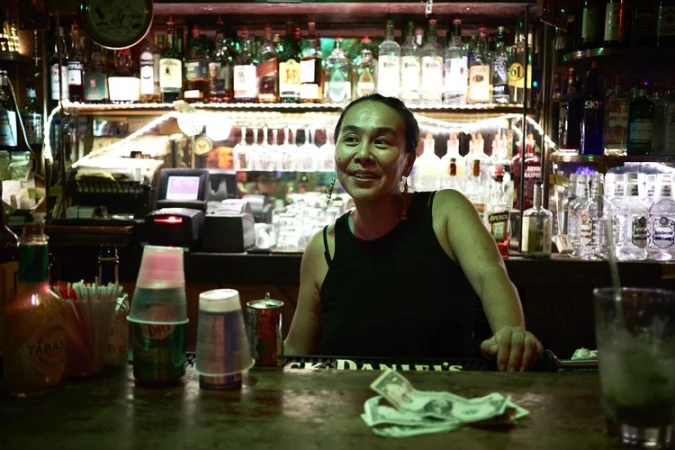 A bartender smiles while standing behind a bar stocked with liquor bottles, glasses, and bar equipment in a dimly lit establishment.