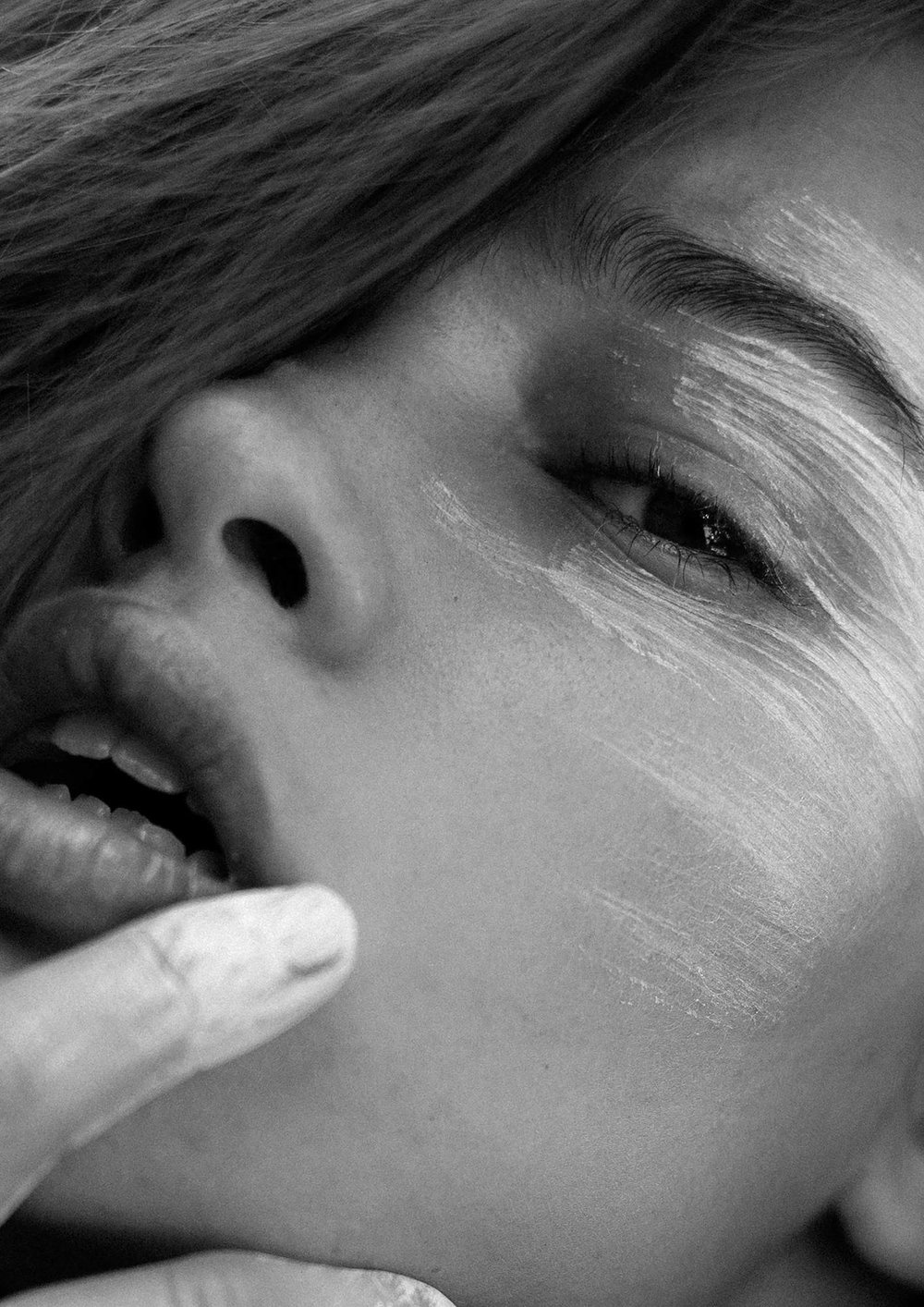 Close-up black and white portrait of a woman with white paint on her face and a finger near her lips in a dimly lit setting.