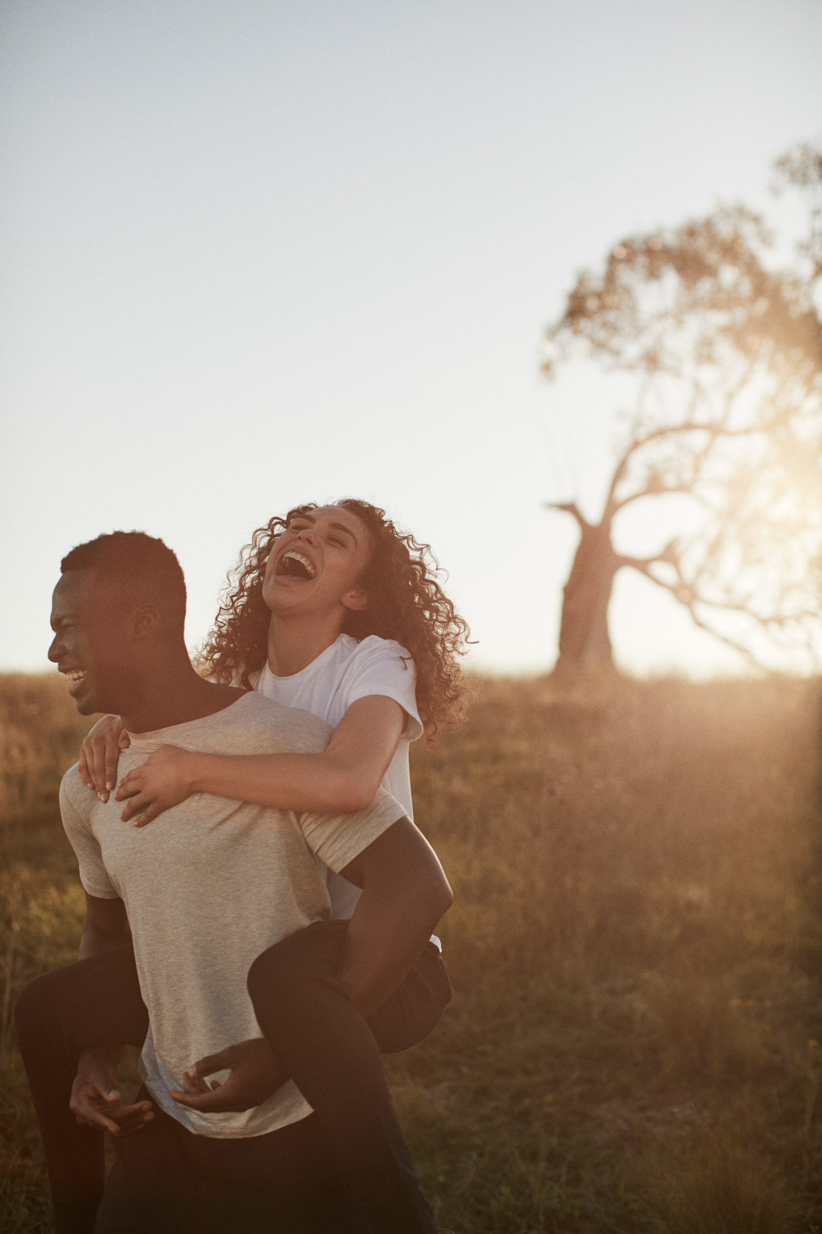 A smiling African-American man gives a curly-haired woman a piggyback ride through a grassy field at sunset, with a tree in the background.