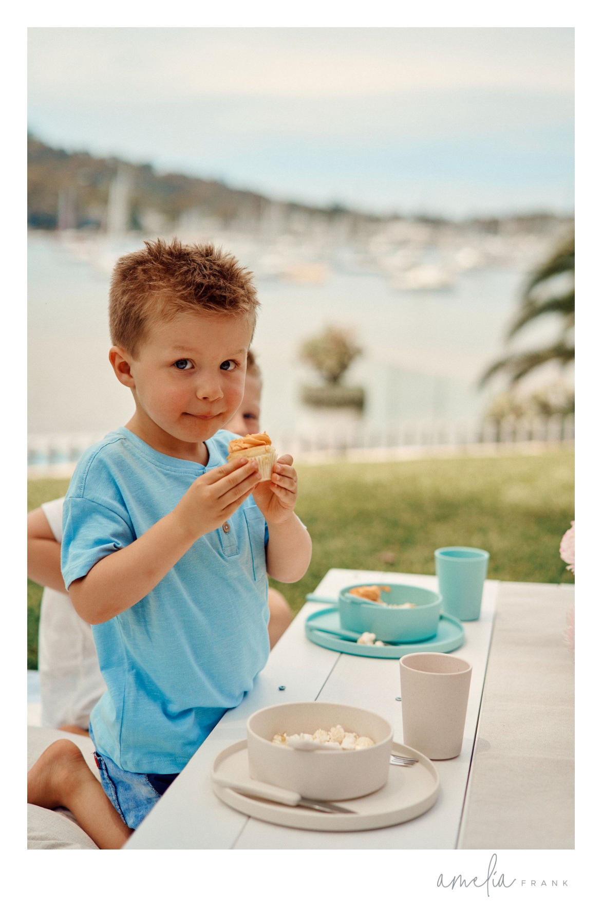 A young boy sits at an outdoor table, eating a cupcake, with dishware visible, and a bay view in the background on a sunny day.