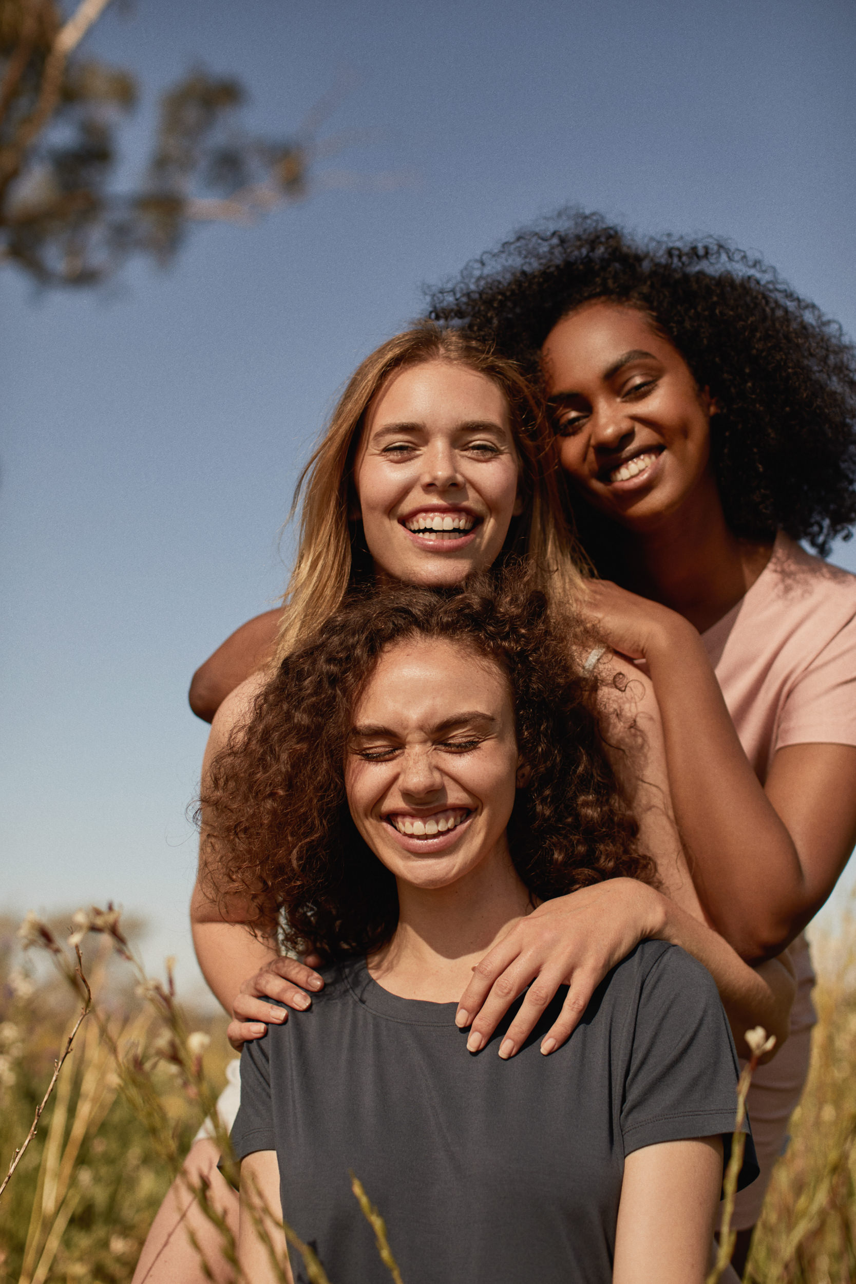 Three diverse women stand in a grassy field, smiling. One has curly hair and wears a dark gray t-shirt, while the others have arms around her.