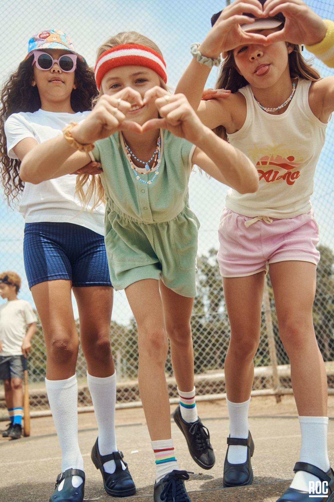 Three young girls in casual clothing stand outside on a sunny day, posing for a photo with heart-shaped hand gestures, smiling.