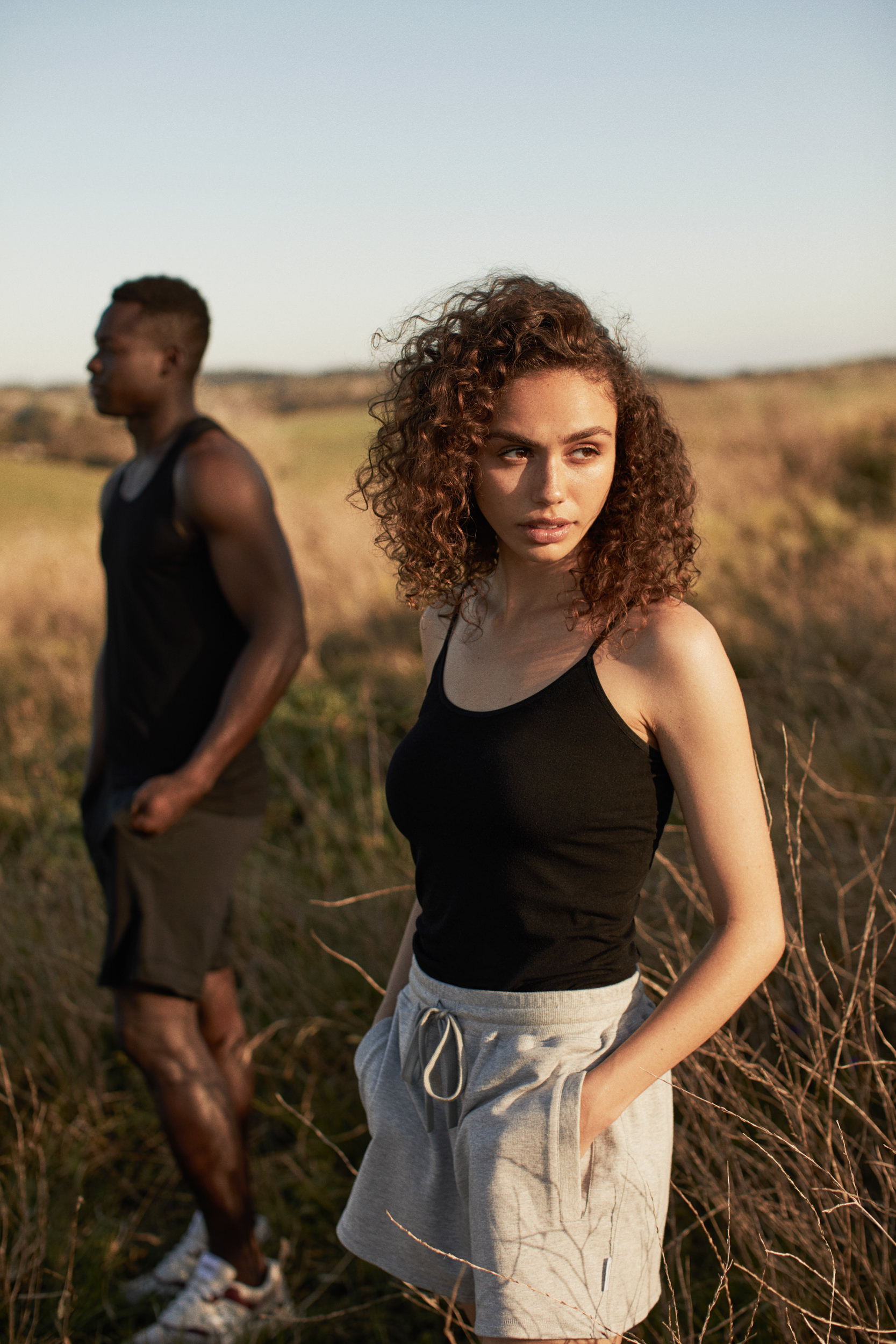Two athletic people in sportswear outside in a grassy field on a sunny day. An adult woman is in focus, with an adult man out of focus.